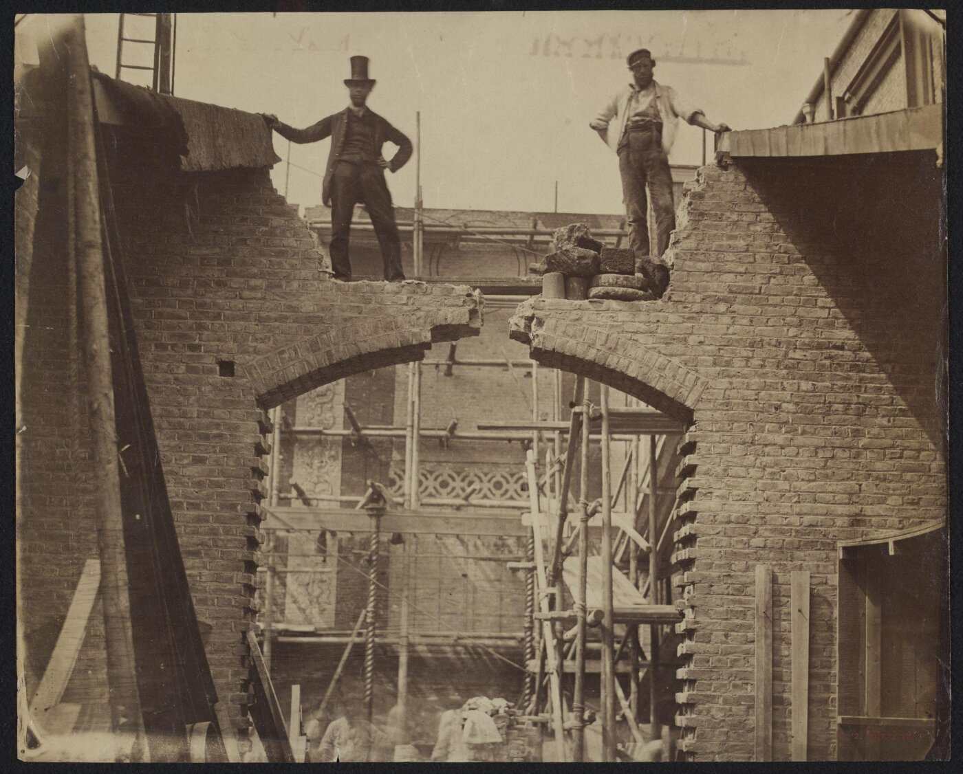 Two men, one wearing a top hat, standing above a brick arch as it's being built